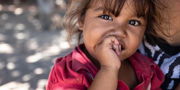 A two-year old girl puts a thumb in her mouth while sitting on her mother’s lap outside on the street. A two-year old girl puts a thumb in her mouth while sitting on her mother’s lap outside on the street.