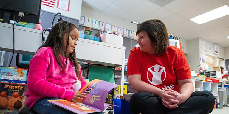 WA, a Save the Children staff member sits next to a little girl in a pink shirt, who's reading a book. WA, a Save the Children staff member sits next to a little girl in a pink shirt, who's reading a book.
