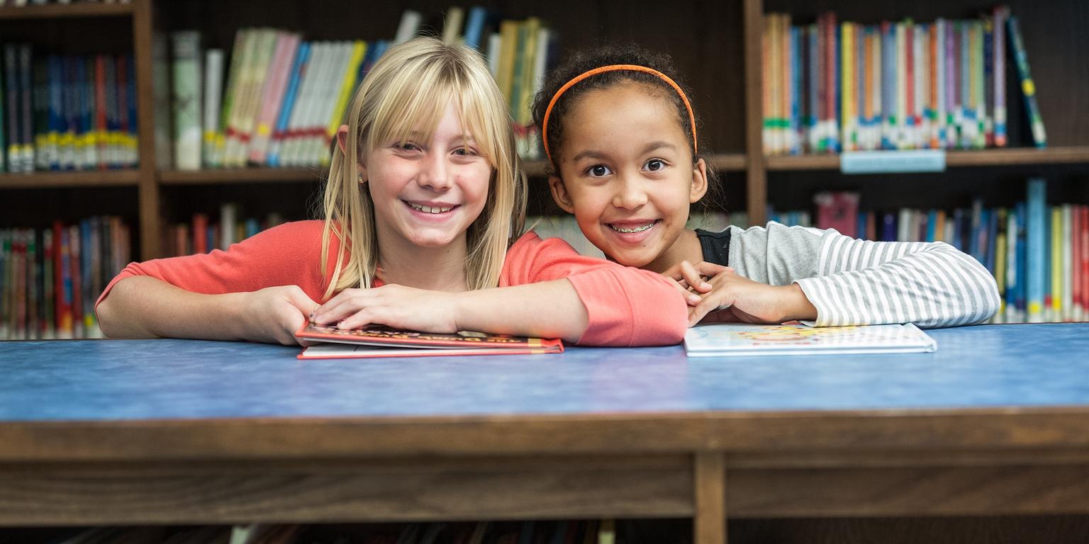 West Viginia, a two girls stand in a library and smile at the camera West Viginia, a two girls stand in a library and smile at the camera