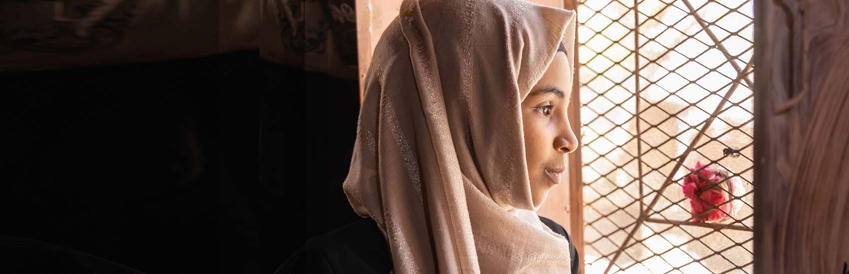 In Yemen, a girl looks out a window while sitting in a dark room. 